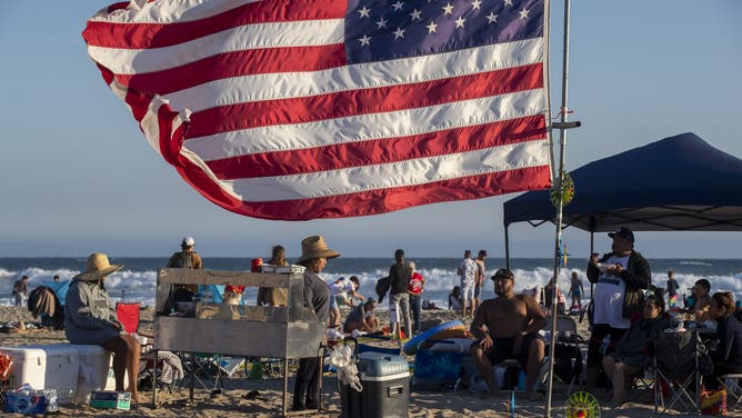 Huntington Beach, CA - July 04: The Espinoza, Vazquez and Romero families of Fallbrook grill tacos on the beach beneath their giant American flag as they await the fireworks over the ocean Sunday, July 4, 2021 in Huntington Beach, CA. The festival featured live entertainment &amp; DJs, merchant &amp; crafts vendor booths, amusements &amp; carnival rides, food trucks, beer &amp; wine garden, sponsor activations, live radio broadcasts, and prize giveaways. A Surf City Run 5K kicked off the day and a neighborhood parade that included 3 separate routes replaced the large parade of years past before the pandemic. The grand finale featured fireworks over the ocean shot off from the pier at 9pm.