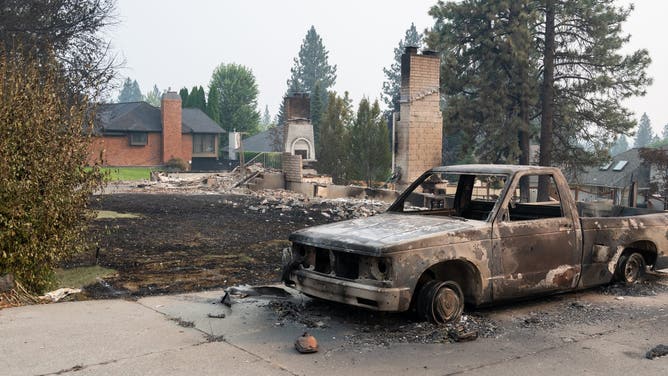 A burned pickup truck sits in the driveway of a burned residential home with an untouched home in the background in the Balboa neighborhood of Spokane, Washington, on August 2, 2026. Fast-moving fires left a trail of destruction in the northwestern US city of Spokane on Sunday, with officials reporting hundreds of structures reduced to cinders. Thousands of people were ordered to evacuate their homes Saturday and overnight into Sunday, as three separate fires broke out in and around the city in the east of Washington state.